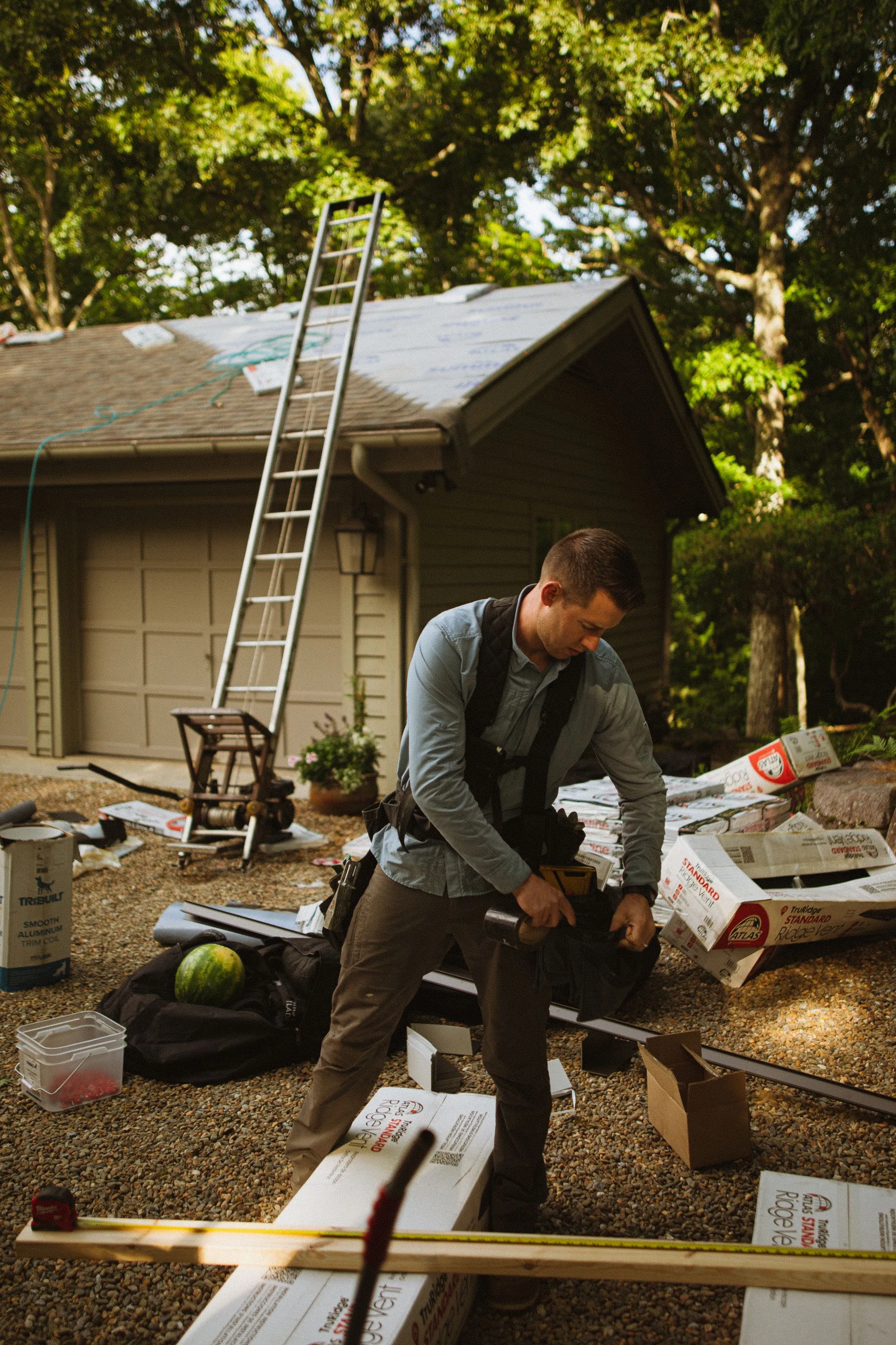 A roofer handling bundles of shingles beside a house under tall trees