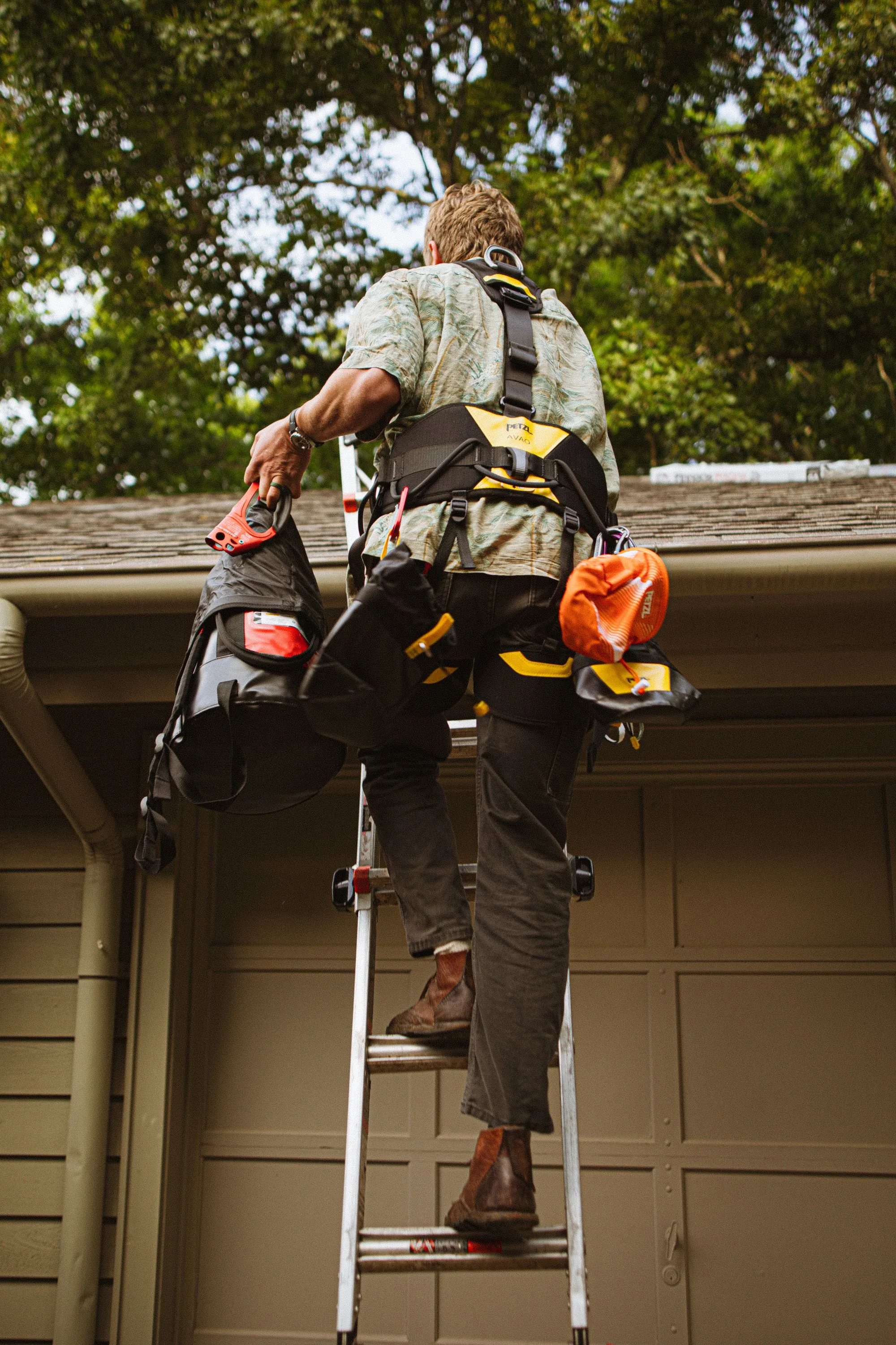 A roofer in a harness climbing a ladder at the eaves of a house